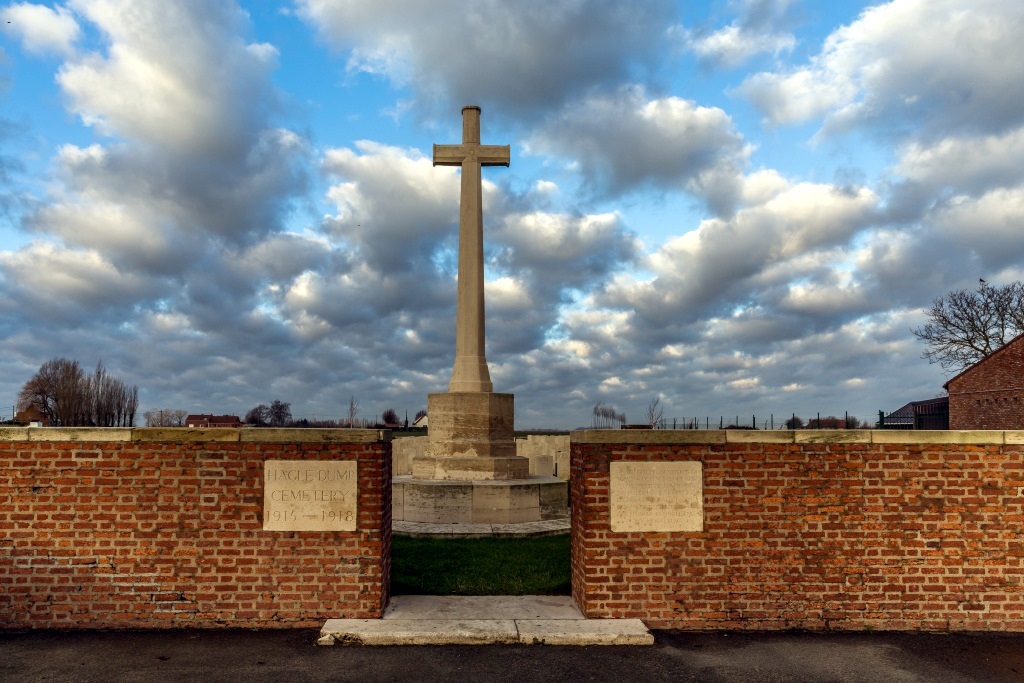 A gap in the brick wall leads to the cemetery and the Cross of Sacrifice is prominent behind the gap