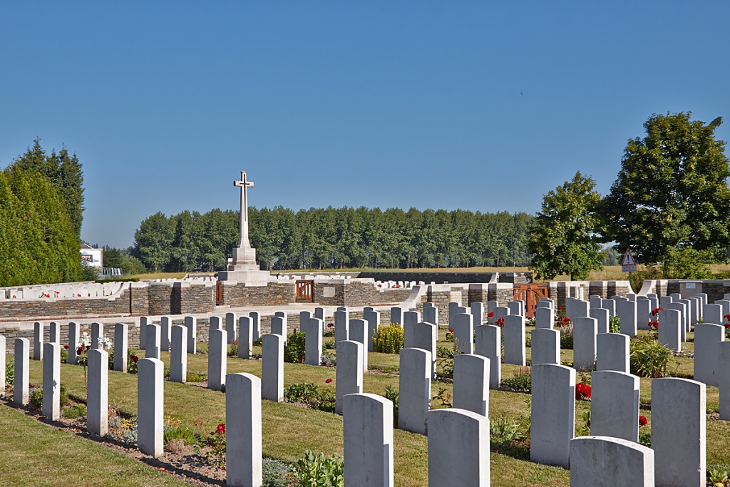 Photo of Sailly-sur-la-Lys Canadian Cemetery. Rows of white headstones in front of 2 walls with more headstones and a white cross monument behind.
