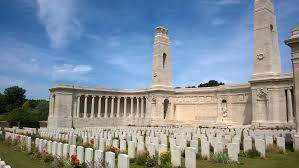 Photo of Vis-en-Artois British Cemetery. Rows of white headstones in front of a stone wall memorial with 2 central towers.