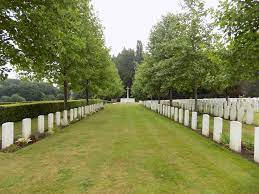 Photo of Kemmel Chateau Military Cemetery. A grass area lined with rows of white headstones surrounded by trees.