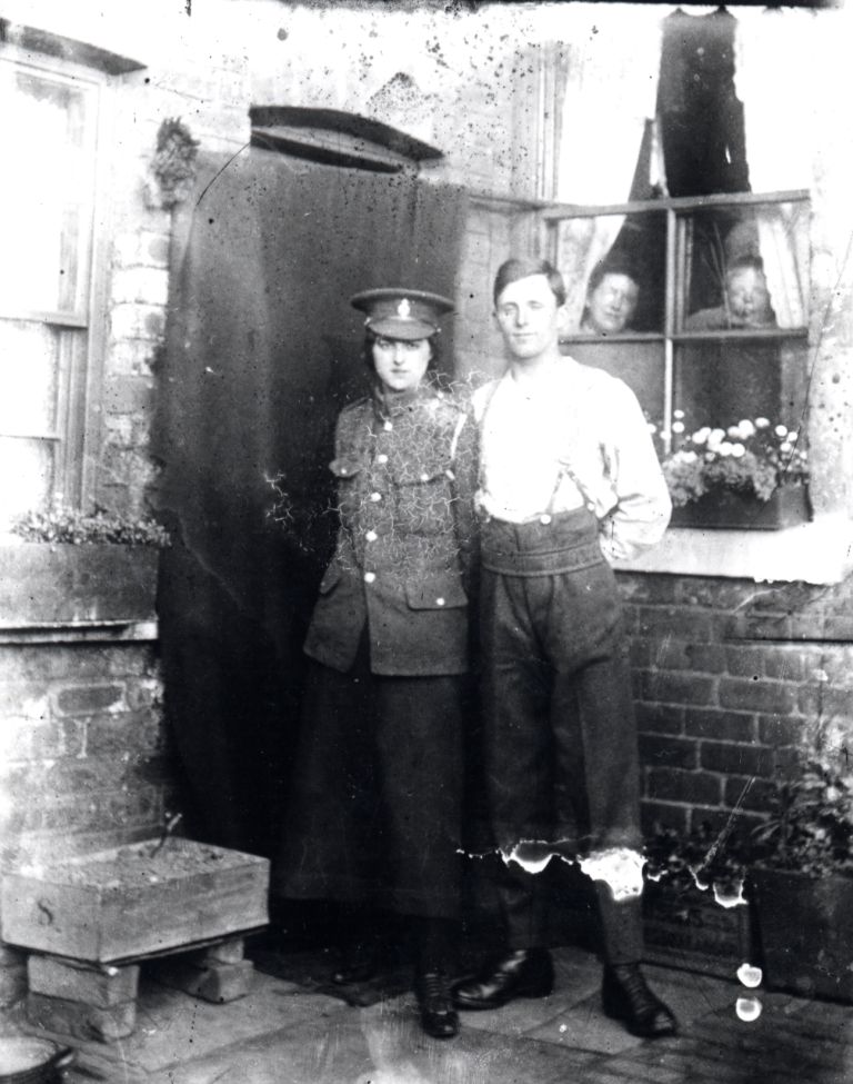 john with his girlfriend. she is wearing his cap and jacket and they are standing outside beside the back door  of a house. two people are looking through a window at them