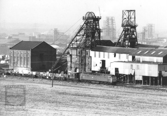 A black and white photograph of Wheldale Colliery, Castleford, in 1984, three years prior to closure.  Two sets of winding gear rise above the colliery buildings.  A train with goods wagons is passing before the pit and in the background is Hicksons and Welch chemical works. The locomotive is a Class 47 (formerly Brush Type 4). Behind the locomotive is a Guards Van, also called a Brake Van. The following vehicles are known as bogie-bolster wagons, used to carry long items such as railways lines or timber. Internal Reference: CA YILLUS 5915a  