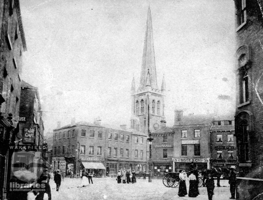 A black and white postcard of a busy day in the  Bullring, Wakefield.  On the left can be seen the Jap Cafe, surrounded by shops.  A horse and cart can be seen on the right, and in the middle of the picture, taken in the early 1900s, can be seen the spire of Wakefield Cathedral, the tallest in Yorkshire.  