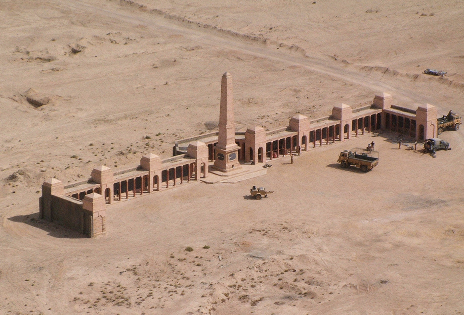 Photo of Basra Military Cemetery. Sand ground with  a long wall memorial with 11 towers. The central tower is the tallest. 3 military vehicles and a trailer are in front,