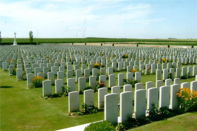 Photo of Bancourt British Cemetery. Rows of white headstones with plants in front of them. A cross monument to the left behind them.