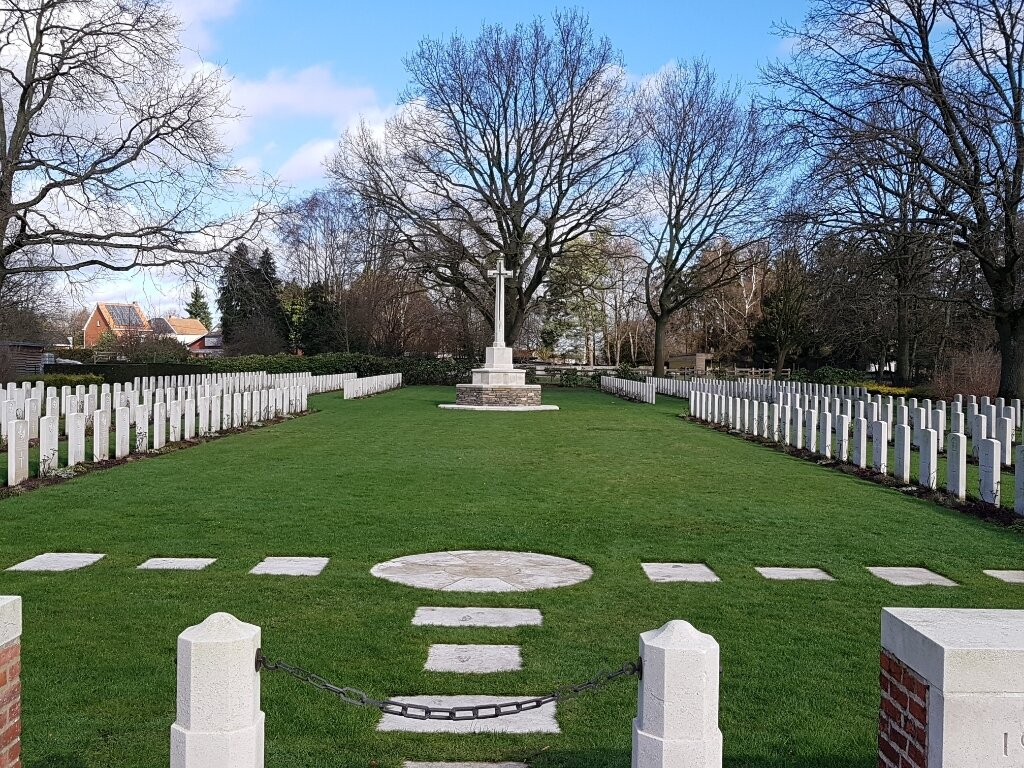 The white Cross of Sacrifice stands at the end of two rows of white gravestones with a large strip of mown grass down the middle. Trees surround the cemetery