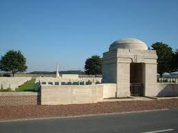 Photo of Gouzeaucourt New British Cemetery. A wall and large square archway in front of a grassed area lined with rows of white headstones.