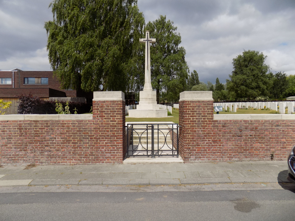 Photo of La Brique Cemetery No 2. A red brick wall in front of a large white stone cross monument and rows of white headstones with trees behind.