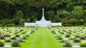 rows of small white graves with a grass path in the middle leading to the white cross of sacrifice in the background. Trees are behind it