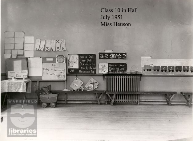 A black and white photograph of a classroom at Snapethorpe Infants School, on the corner of St George's Road and Broadway, Lupset, in July 1951.  The work of class 10, taught by Miss Henson, is displayed on the wall, the result of English lessons.  A picture of a train is also shown.  Benches and a toy pram lie against the wall.  The school, established in 1931, later became part of Snapethorpe Primary School. Internal Reference: Private Collection