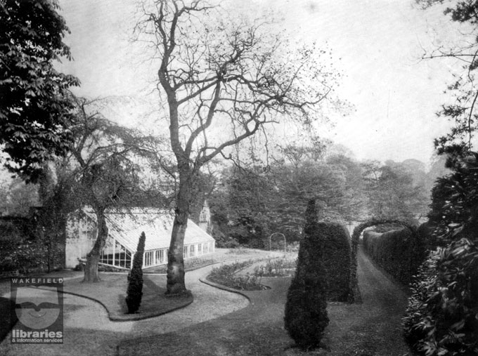 A black and white photograph of the rose garden at Kettlethorpe Hall, Wakefield.  The photograph is taken from the sales brochure of 1908 when the estate was sold at auction.  The view is from the dining room and shows winding paths, lawns with rose flower beds, hedges and archways.  To the left is a large greenhouse and beyond the garden are many large trees.  Internal Reference: WR (XH)