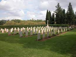 rows of white gravestones surrounded by mown grass. around the perimeter is a hedge and trees and the white cross of sacrifice is at the far end