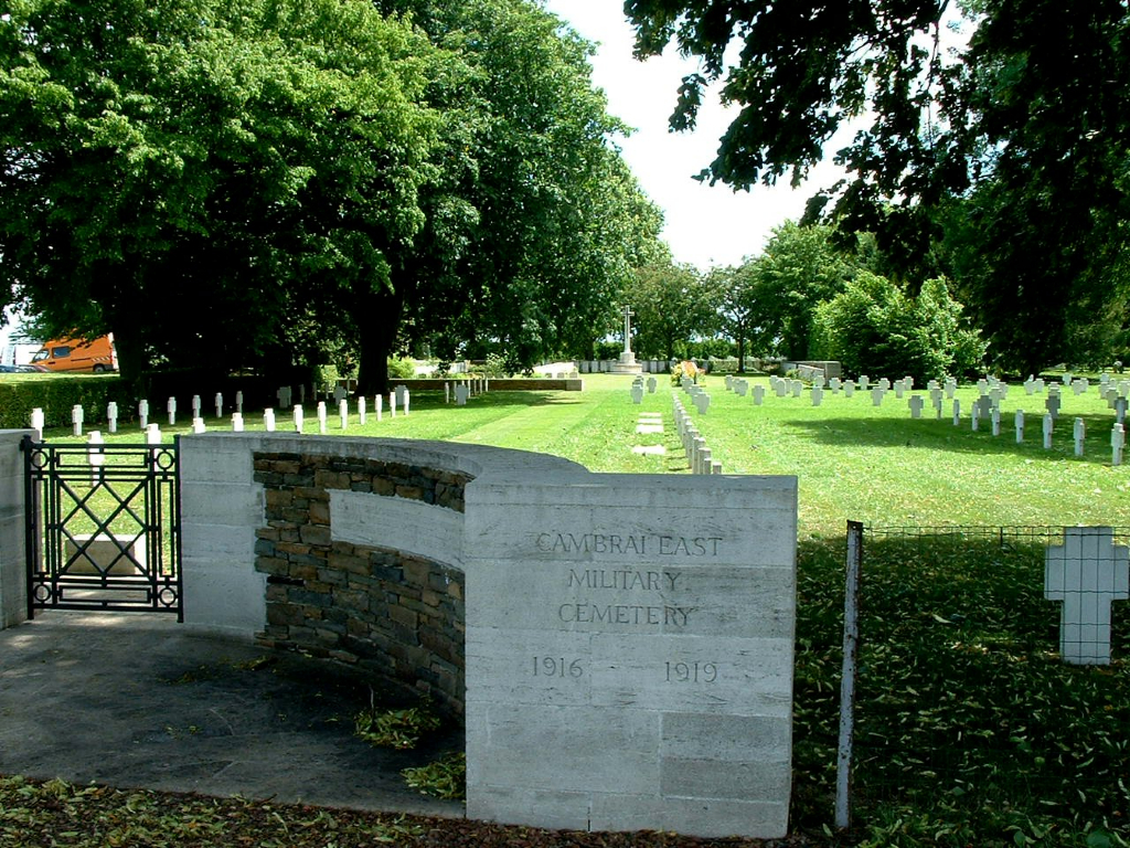 the stone entrance to the cemetery which is surrounded by trees. the rows of graves stretching into the distance can be seen surrounded by mown grass
