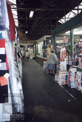 A colour photograph of the goods part of the indoor market at Castleford, in 1991.  Clothing stalls are to the left and rear of the photograph.  On the right is a stall selling toiletries and cleaning products.  Shoppers are visiting the market.  Internal Reference: CA YILLUS 3544