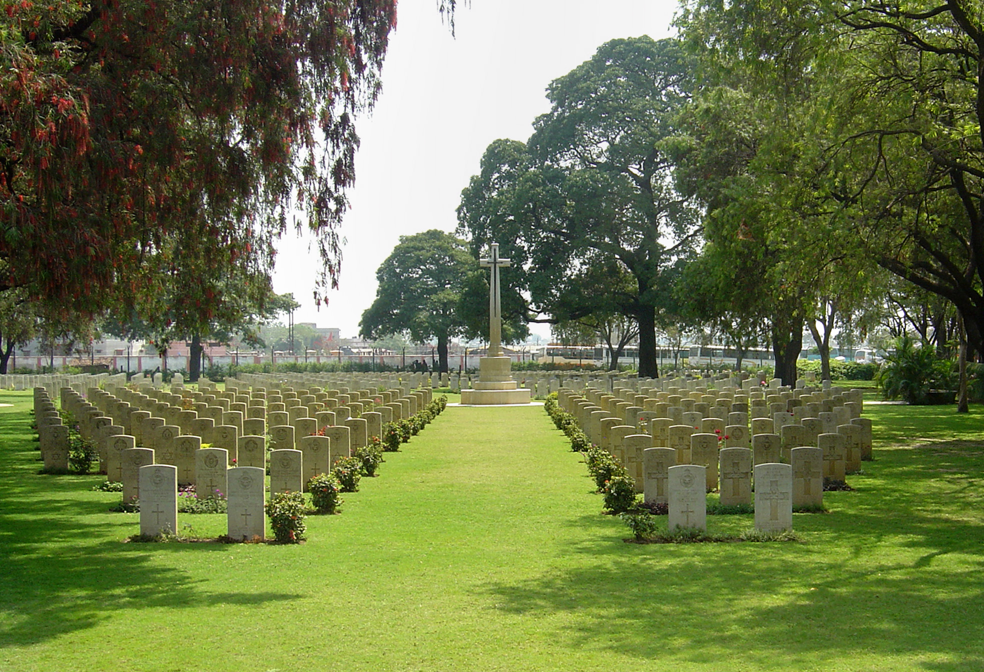 rows of graves with a wide expanse of grass between the 2 blocks of gravestones leads to the white cross of sacrifice at the far end of the cemetery. Several trees surround the cemetery