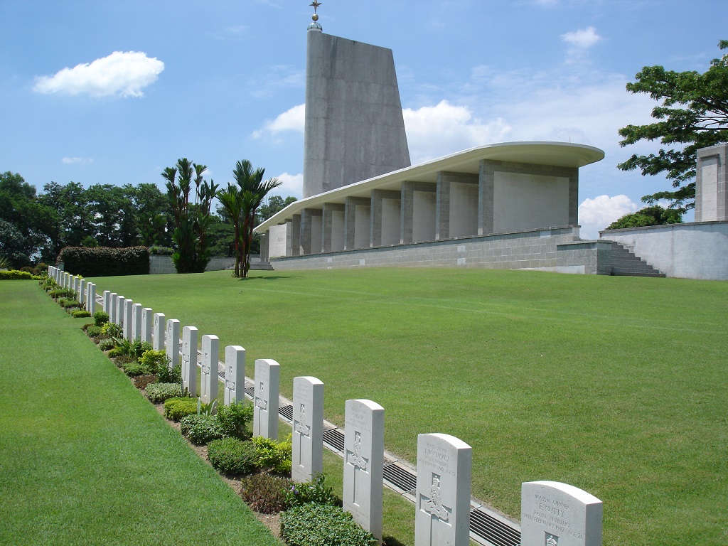 Photo of Singapore Memorial. Grass area with a line of white headstones with plants in front. A large grey stone monument with a tower behind.