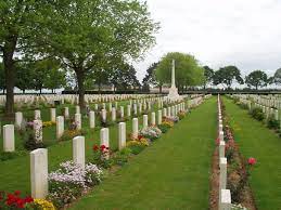 Rows of white gravestones with plants flowering in front of them and mown grass between the rows lead to the white Cross of Sacrifice at the far end. Trees surround the cemetery