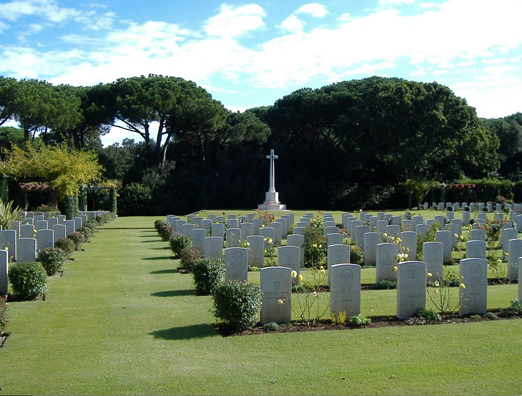 Photo of Beach Head Memorial Anzio. Rows of white headstones in front of a cross memorial with a background of trees.