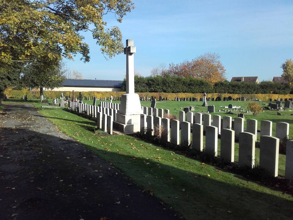 a gravel path on the left with an overhanging tree. the white cross of sacrifice and two rows of white gravestones on the right