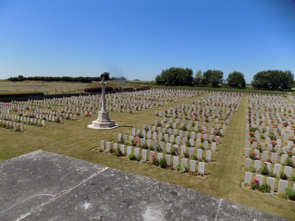 Looking down into a graveyard with rows of white gravestones and plants in front of them. Areas of mown grass are between the rows and the white Cross of Sacrifice can be seen