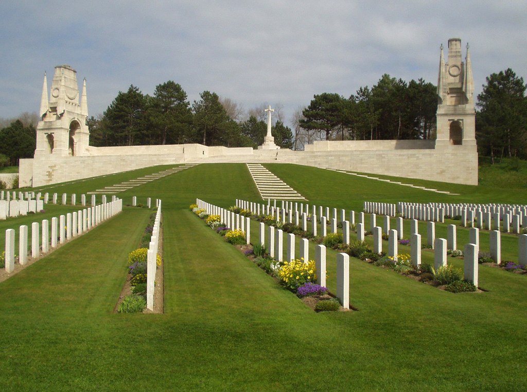 Photo of Etaples Military Cemetery. Rows of white headstones with a large memorial with towers either side and a cross in the middle.
