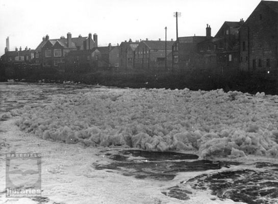 A black and white photograph taken from Lock Lane looking across to Aire Street, Castleford showing foam pollution on the River Aire.  Detergent from nearby textile mills washed down the river and was churned up when passing over the weir.  Internal Reference: CA YILLUS 1089