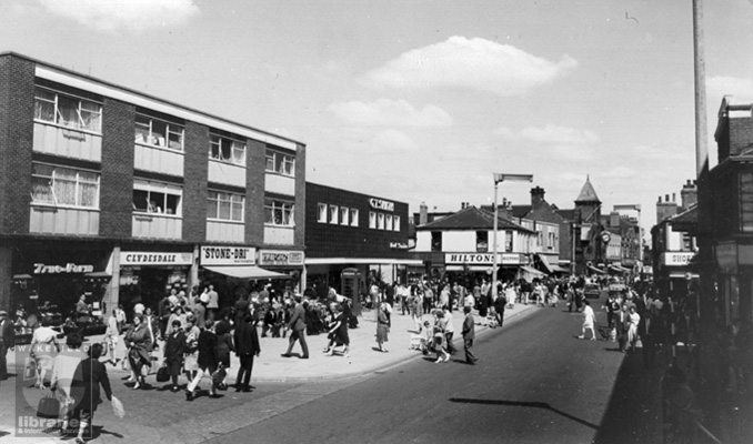 A black and white photograph of Carlton Street, Castleford taken from near Marks and Spencer's shop looking towards Bridge Street in 1965. It is a busy shopping day and on the left hand side of the photograph the shops include True-form, Clydesdale, Stone-dri, Halfords, G T Smith, Hilton and the Market Hall. On the right hand side of the street the shops can only be seen in profile and no shop names are visible. Internal Reference: CA YILLUS 750