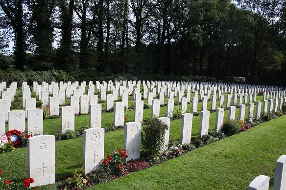 Photo of Oosterbeek Military Cemetery, Arnhem. Rows of white headstones with flowers and plants in front of them. A row of trees in the background.