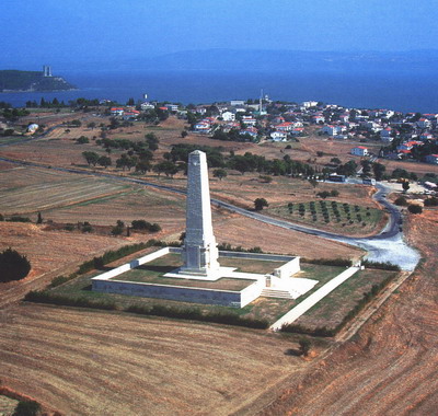 Photo of Helles Memorial. Large white stone tower bordered by a square white stone wall surrounded by fields with a town behind.