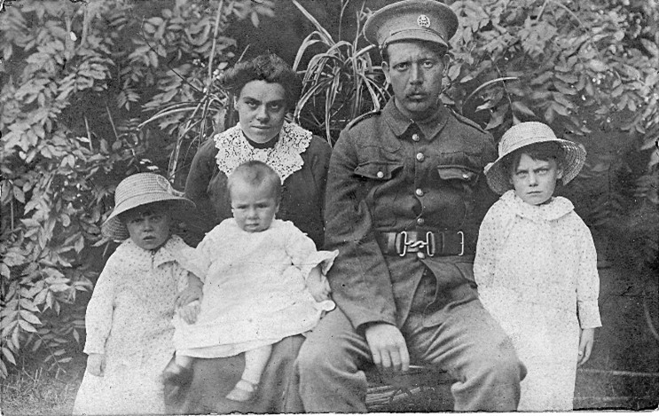 aaron smeaton with his family. they are all seated, his wife sits to the left of him with a baby on her lap and a young girl stands beside her, another young girl stands next to aaron, he is wearing his army uniform and has his army cap on