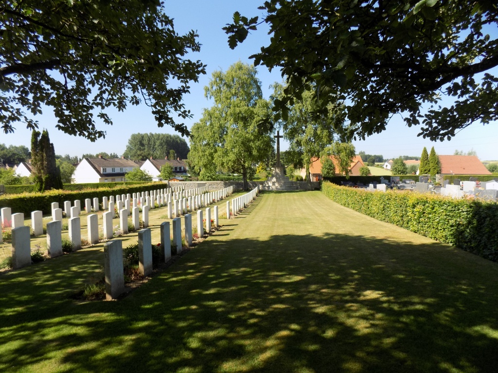 Four rows of white gravestones partly in the shade from overhanging trees are within an area of mown grass surrounded by a hedge. Buildings of a town are in the distance