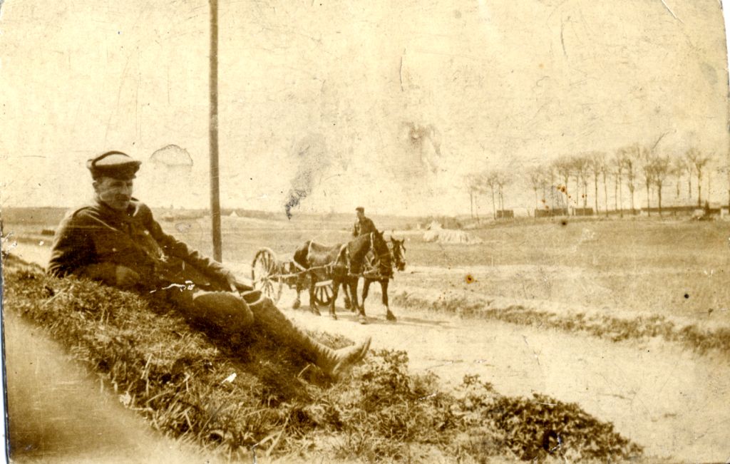 sepia photograph of a man sitting beside a road with a horse and cart in the distance. Taken at the Front