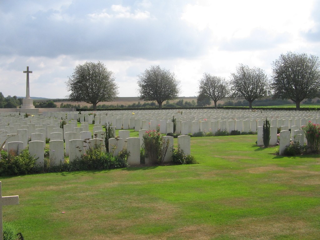 Photo of Aubigny Communal Cemetery. Rows of headstones in front of a cross memorial and a line of trees.