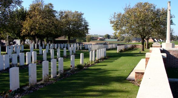 Photo of Henin Communcal Cemetery. Rows of headstones surrounded by trees with a side view of a cross memorial to the right.