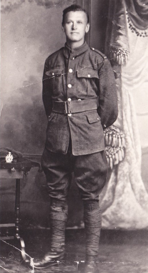 john richard wood stands proudly by a table which has his army cap on it, he is wearing his army uniform the photograph was probably taken in a photographic studio as there is a painted background and a heavy curtain to one side