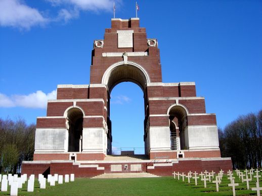Photo of Thiepval Memorial. Red brick and white stone memorial with 3 arches. Rows of headstones to the left front. Cross markers to the right front.