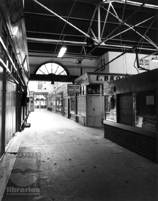 A black and white photograph of the section of Castleford Indoor Market which led out onto Carlton Street.  The market is closed and the stalls are shuttered.  James, upholsterers, Bromley and Vairy, Drapers and a herbalist are amongst those in the picture.  Internal Reference: CA YILLUS 686