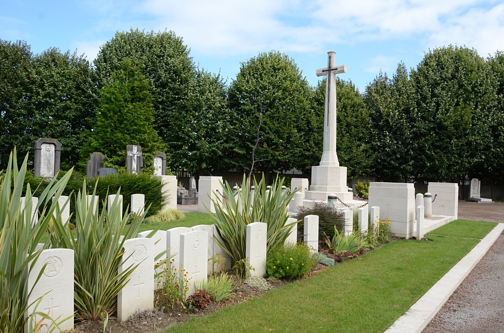 Photo of St Andre Communal Cemetery. Double rows of white headstones and plants in front of a cross memorial with trees behind.