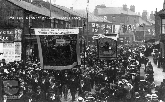 A black and white photograph of a miners' demonstration march along Aire Street, Castleford in 1903.  Three banners are being carried saying 'Wheldale and Fryston - 36 Weeks dispute', although the dispute laster 68 weeks, spanning 3 years.  A brass band marches amongst the miners and the street is lined with onlookers.  Internal Reference: CA YILLUS 3706