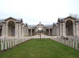 Photo of Arras Memorial. Rows of headstones with a central grass path in front of a stone building with 2 archways and columns.
