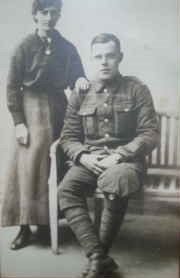john williamson is seated on a wooden bench, he is wearing his army uniform and his wife stands beside him