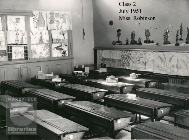 A black and white photograph of a classroom at Snapethorpe Infants School, Broadway, Lupset, in July 1951.  The picture shows the empty desks and school work of the children of class 2 taught by Miss Robinson.  Paintings of people are displayed on the left, and of trees ad other plants on the right.  On some of the desks are models, a train, a tank and a ship made of boxes.  The school, established in 1931, became part of Snapethorpe Primary School in 2003. Internal Reference: Private Collection