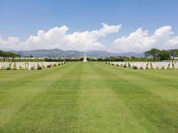 Wide expanse of grass with white gravestones to either side, leading up to the white cross of sacrifice in the background. further in the background mountains can be seen