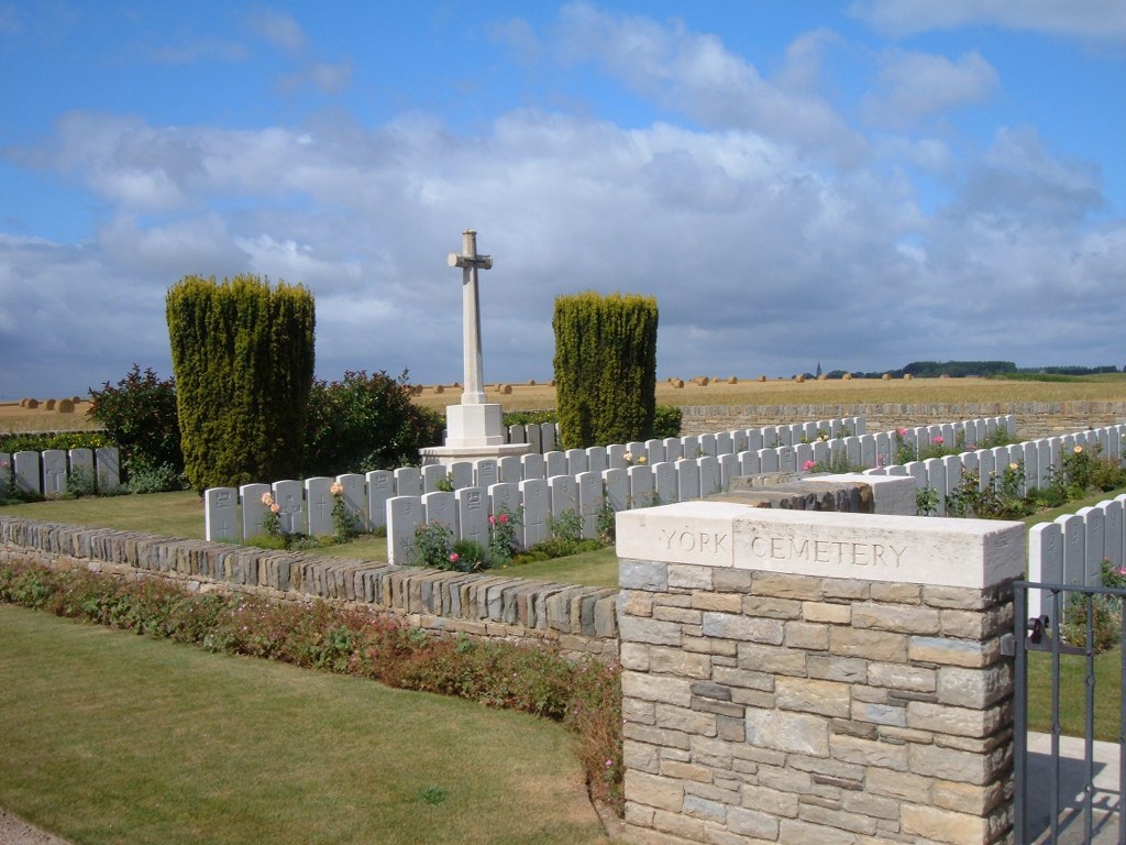Photo of York Cemetery Haspres. A stone wall with York cemetery inscribed on the top in front of rows of white headstones that surround a large white stone cross monument .