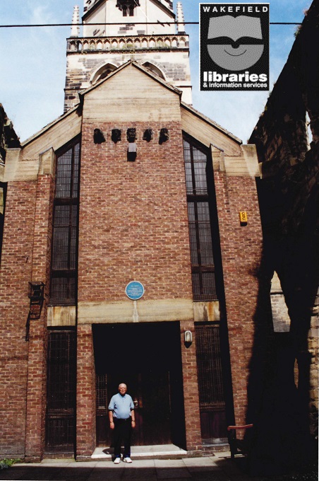 A colour photograph of the modern entrance to All Saints Church, Potnefract. Local historian Mr R Gosney is standing in front. The blue plaque can be seen above him. Internal Ref: PO
