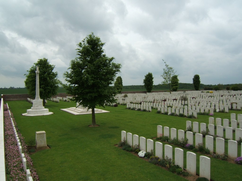 Rows of white gravestones in a cemetery with mown grass. nearby is the Cross of Sacrifice and a tree