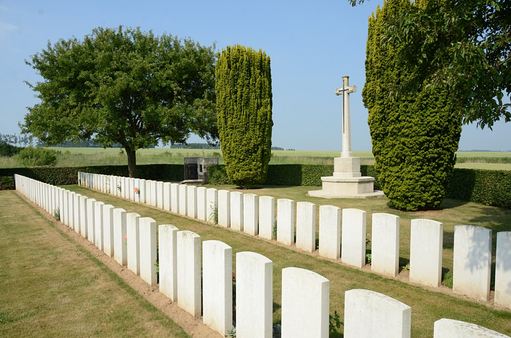 Two rows of white gravestones with mown grass in between the rows. The white Cross of Sacrifice stands behind them. The graveyard is surrounded by a hedge and some trees are nearby